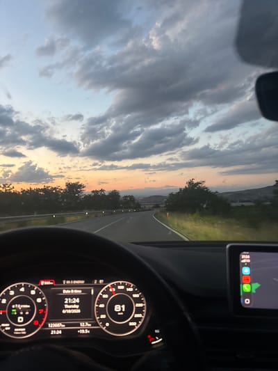The image shows a view from the driver's seat of a car on a road near Aleko Konstantinovo, Bulgaria, during sunset. The sky is filled with dramatic clouds, with hues of orange and pink from the setting sun. The road curves ahead, bordered by a guardrail and roadside lights. Trees and rolling hills form the landscape on either side of the road. The car's dashboard is visible, showing the speedometer at 81 km/h, the date and time as 21:24, Thursday, 6/18/2026, and odometer reading 123687 km. A central touchscreen displays a navigation interface. The scene captures a journey through a rural landscape at dusk.
