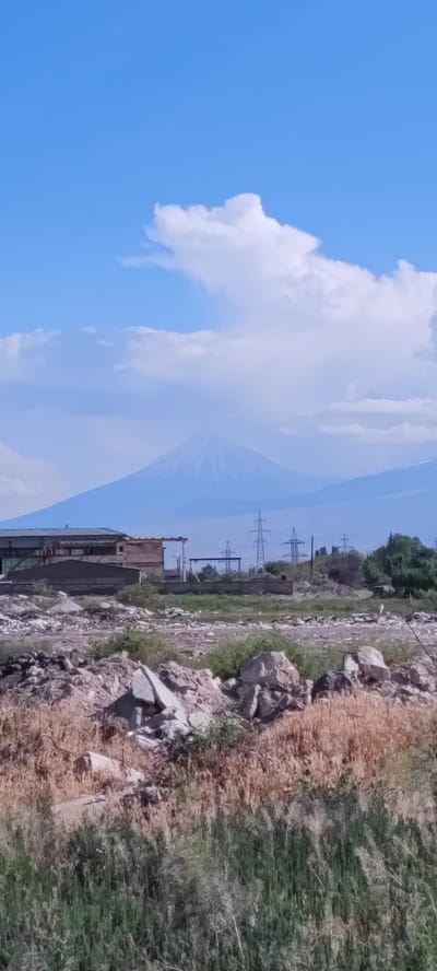 The image displays a landscape in Ayntap, Armenia, under a bright blue sky with scattered white cumulus clouds. In the distant background, a large, snow-capped or light-colored mountain, identifiable as Mount Ararat, rises prominently, partially obscured by atmospheric haze and lighter clouds. To its right, additional hazy mountain ranges are visible.

The midground features a semi-developed or industrial-agricultural area. On the left, a low, rectangular building with a dark roof and light-colored walls is visible. Extending horizontally across the midground in front of the building is a low, concrete-like wall or barrier. To the right of the building, a series of metal lattice electricity pylons stand against the mountainous backdrop. The ground in this section consists of undeveloped land, characterized by patches of green shrubbery, dry, light brown grasses, and significant amounts of scattered light-colored rocks and rubble.

The foreground is dominated by uncultivated ground. It is covered with a mix of dry, light brown grasses and thicker, darker green scrub vegetation. Numerous broken rocks and fragments of debris are strewn across the immediate ground plane. No people or animals are discernible in the scene, and no specific actions or interactions are depicted. The environment suggests a rural or peri-urban setting with both natural and industrial elements.