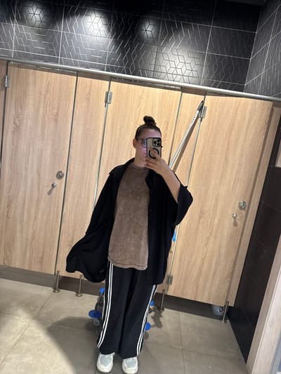A person is standing in a restroom in Gil'deevo, Russia, taking a selfie in front of a row of wooden stall doors. The person has dark hair pulled back in a bun and is wearing glasses, a brown shirt, a black cardigan, and black sweatpants with white stripes. They are holding a cell phone up to their face to take the picture. The restroom has dark gray tile on the wall and light gray tile on the floor. The stall doors are light wood with metal hardware.