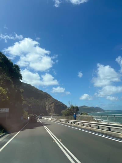 A road spans the frame, bordered by a barrier and a mountain. The ocean and more mountains are visible beyond. A blue-clad person stands near the barrier, adding a human element to the landscape. The road surface is marked with white lines. Clouds scatter across the expansive blue sky. The scene is bathed in daylight, with shadows cast along the road, suggesting the time of day. A sign near the roadside provides some context.