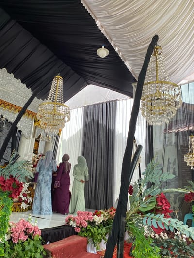 This image depicts an elaborately decorated event space, likely an indoor or tented venue, located in Geulanggangbaro, Indonesia. The overhead structure features a draped fabric ceiling, segmented into pleated black sections on the left and white sections on the right, forming a canopy. A single white domed light fixture is suspended from the white ceiling section.
Multiple chandeliers with gold-toned frames and numerous clear crystal teardrop embellishments are suspended, with one prominently in the midground and another partially visible on the far right. Thick, black fabric-wrapped poles or columns ascend from the foreground, providing structural support; one pole is positioned directly in front of the central chandelier.
A raised, glossy white stage occupies the midground. Its backdrop consists of intricate, carved white panels adorned with gold and black decorative accents, featuring scrolled motifs and repeating circular patterns. Red and white floral arrangements are placed at the base of the backdrop on the stage.
Three adult females, viewed from the back, are standing on the stage, facing the decorative backdrop. The woman on the left wears a light blue long-sleeved dress with a floral pattern and a grey hijab. The central woman is dressed in a dark maroon or purple long-sleeved dress and a brown hijab. The woman on the right wears a light sage green long-sleeved dress and a matching light green hijab, with her hands clasped behind her back.
The immediate foreground is extensively decorated with floral arrangements, including clusters of pink roses, deep red hydrangea-like flowers, and various green foliage, some with silver-green, fern-like leaves. A low, rectangular object with a red, textured surface (possibly upholstered wood) is partially visible in the bottom right foreground.
The background beyond the stage is enclosed by floor-to-ceiling drapes, featuring sheer white panels alternating with dark grey or black opaque panels. Through the sheer drapes on the far right, a faint outline of what appears to be a brick wall or an exterior building structure is discernible.