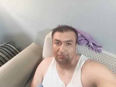 A man is seen from the chest up, looking at the camera. He is wearing a white tank top. He has short, dark hair, brown eyes, and stubble on his face. He is sitting on a couch, with a patterned cushion behind him and a purple blanket draped over the back of the couch. The background appears to be a textured wall. The lighting suggests an indoor setting, possibly a living room. No specific location in Ashgabat, Turkmenistan can be determined from the image.
