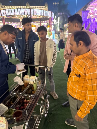 At an outdoor event, a food service worker wearing gloves prepares food from a cart. Several men stand nearby, including one in an orange checkered shirt and another in a pink shirt. Background shows an illuminated structure.
