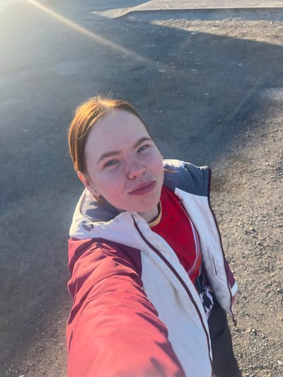 A person is standing outdoors on a paved surface, likely asphalt or concrete, in direct sunlight. The individual is wearing a red patterned shirt beneath a partially unzipped jacket with white, gray, and maroon panels. The person is fair-skinned with light hair that has some sunlit highlights. Their face is tilted upward towards the camera. Shadows cast by nearby structures are visible on the left side of the image. There is no discernible information in the image to determine the location in Nizhnie Yaki, Russia.