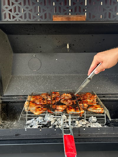 A close-up, eye-level shot shows a person's right hand holding a silver-handled knife, with the blade pointed towards a grill basket filled with glistening, glazed chicken pieces over hot coals. The grill basket is positioned inside a dark, black metal barbecue. The barbecue has a decorative metal grate with a geometric pattern on the top back wall, and a wooden block rests on it. The environment appears to be outdoors, given the natural light and the context of a barbecue. There is no discernible information about the specific location within Starokorsunskaya, Russia, or any other identifying features that would narrow down the precise setting beyond it being a barbecue area. The focus is on the act of preparing food, with the chicken appearing to be marinated or basted with a dark sauce. The coals beneath the grill are visible, covered in white ash, indicating they are hot and actively producing heat. The overall scene suggests a culinary activity, likely a meal preparation.