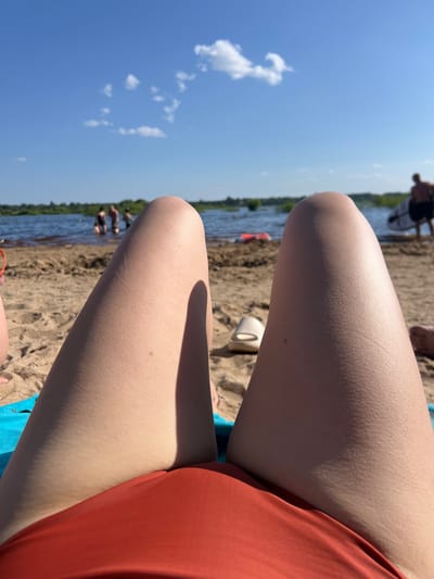 The image shows a first-person perspective of someone lying on a sandy beach, facing a body of water under a clear blue sky with scattered white clouds. The foreground is dominated by the person's thighs and lower torso, wearing an orange garment, likely a swimsuit bottom. To the left and right of the thighs are exposed legs. The beach sand is visible in the foreground and mid-ground. In the mid-ground, there are several people in the shallow water, some appearing to be swimming or wading. Further back, on the right side of the image, a person stands near the water's edge holding a surfboard or paddleboard. The background consists of a tree line along the edge of the water, suggesting a natural landscape. The overall scene depicts a recreational day at a beach, possibly in a natural area outside of a densely populated urban environment. The location is specified as Ust'ye, Russia, implying a riverside or lakeside beach setting.