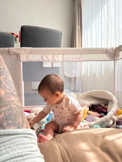 The image shows a baby playing inside a playpen filled with various toys. The baby is wearing a light-colored onesie with a strawberry pattern. The playpen has mesh sides and a light pink frame.
In the background, there are large windows with white blinds, suggesting it is daytime. To the left, a portion of a grey upholstered chair is visible. To the right, a person's legs, covered in tattoos, are in the foreground, partially obscuring the playpen. A large, light-colored fabric item, possibly a couch or cushion, is also visible on the right side of the frame.
The overall scene appears to be a domestic indoor setting. The lighting suggests natural light, possibly late morning or early afternoon. The baby's posture and engagement with the toys indicate active play. No visible text is present in the image. The location context is Veghel, Netherlands.