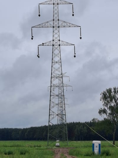 The image shows a large, lattice-style electrical pylon standing tall against a cloudy sky. The pylon is made of metal and has a complex geometric structure. Several crossbars extend from the main structure, each holding a large, spherical insulator. The pylon dominates the left side of the frame, angled from bottom left to top right.
To the right of the pylon, a dense, dark green forest forms a backdrop, with trees reaching towards the sky. A bright green, grassy field runs along the right edge of the image, leading the eye towards the background. In the far right of the image, a small, white and blue portable toilet is visible in the grassy area, near the edge of the forest. A thin, light-colored rope or cable stretches diagonally from the upper right corner of the frame down towards the middle right, disappearing behind the pylon.
The sky is overcast, suggesting a cloudy or potentially rainy day. The overall lighting is diffused. The image appears to be taken outdoors in a rural or semi-rural setting, likely during daylight hours given the overall illumination. The presence of the electrical pylon suggests a power transmission line. The location context provided is Lubnica, Poland. There is no discernible activity or event taking place, other than the static presence of these elements. No text is visible in the image.