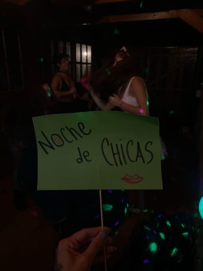 The image shows a dimly lit indoor scene, likely at a party or gathering. Two women are visible in the background. A green sign is held up, displaying the words "Noche de Chicas" (Girls' Night) in black marker, with a lipstick drawing near the word "Chicas." A hand holding the sign is visible on the right side of the frame. The scene is illuminated by green and blue light effects. The location is possibly in Monte Grande, Argentina.