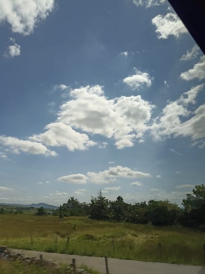 The image depicts a scenic view of a rural landscape under a partly cloudy sky. The foreground features a grassy field with scattered wooden posts and a low stone wall. In the middle ground, a dense line of trees, including some evergreens, creates a natural border. Beyond the trees, rolling hills fade into the distance. The sky is a vibrant blue, dotted with fluffy white cumulus clouds, some of which are illuminated by sunlight. The overall impression is one of a peaceful, natural environment, likely captured from a moving vehicle given the slight motion blur in the foreground and the angle of the shot, suggesting a road trip through the countryside. The location is identified as Burtonport, Ireland, a coastal village known for its natural beauty.