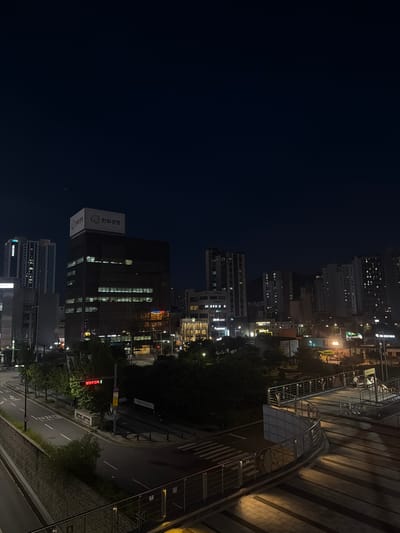 The image depicts an urban nighttime scene in Uijeongbu-si, South Korea. In the foreground, a curved walkway with metal railings is illuminated by artificial lights, casting shadows on the paved ground. Beyond this, a street with crosswalks and traffic lights is visible, with a few trees lining the roadside. A dark, multi-story building with illuminated windows stands prominently on the left side of the frame, displaying a sign that reads "한화생명" (Hanwha Life Insurance). In the background, a cluster of taller residential buildings with numerous lit windows stretches across the horizon, suggesting a densely populated area. The overall atmosphere is that of a quiet, nocturnal city environment.