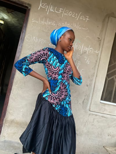 A young woman wearing a blue headscarf, a colorful blue, pink, and black patterned blouse, and a black skirt poses against a textured gray wall with chalked inscriptions. The inscriptions include strings of numbers and letters such as "KG/LLF/2025/807" and "103/101/57-60." The woman is touching her face with one hand and has the other resting on her hip. A window is visible near the top of the wall and a doorway is partially visible in the bottom left corner.