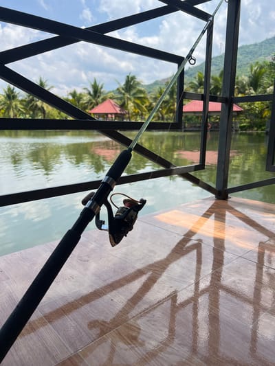 In Suối Tân, Vietnam, a fishing rod with a reel is positioned in the foreground, its black handle and the metallic reel clearly visible. The rod extends diagonally upwards and to the right, crossing over a body of water. In the background, palm trees line the shore of the water, interspersed with two red-roofed structures. Beyond the trees, a lush green mountain rises against a bright blue sky dotted with white clouds. The scene appears to be on a wooden deck or balcony, with sunlight casting geometric shadows of a metal railing onto the floor.