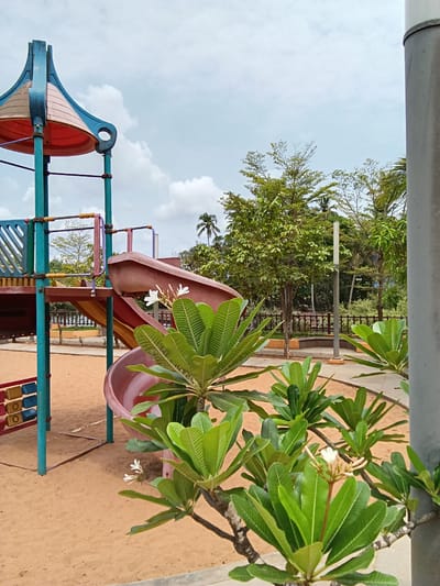 The image shows a playground scene in Chopdem, India. A brightly colored playground structure, including slides and climbing apparatus, is set on a sandy surface. Green trees and vegetation are visible in the background, partially obscured by a metal fence. In the foreground, branches with lush green leaves and white flowers frame the scene. The sky above is pale blue with scattered clouds. A grey pipe runs along the top edge of the frame.
