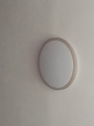 A circular, frosted white light fixture with a beige trim is mounted on a plain, off-white ceiling. The light fixture is centrally located in the frame and is emitting a soft, diffused light. The ceiling appears to be a standard residential or commercial ceiling with a slightly textured surface. There are no other objects or people visible in the image. The overall impression is one of a simple, functional light source in an indoor environment. This image is not specific to the city of Gerbi, Russia, and does not contain any information to suggest its location within that city.