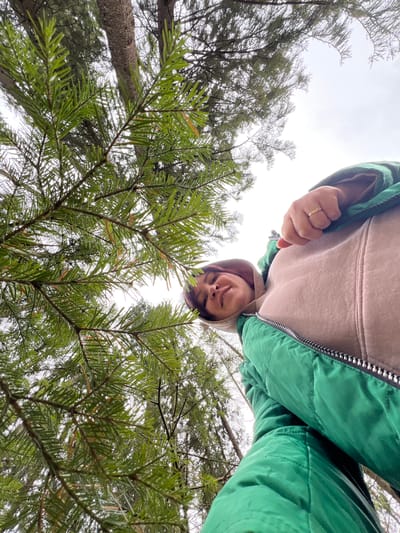 A low-angle, selfie-style photograph shows a person standing upside down in a forest or wooded area. The person is wearing a beige hooded sweatshirt under a green puffy jacket with a silver zipper. A gold ring is visible on a finger. The trees, predominantly evergreens, fill the lower half of the image and frame the person's face. The background shows a light grey or overcast sky. The location is not evident in the image to confirm it is Bol'shakino, Russia.