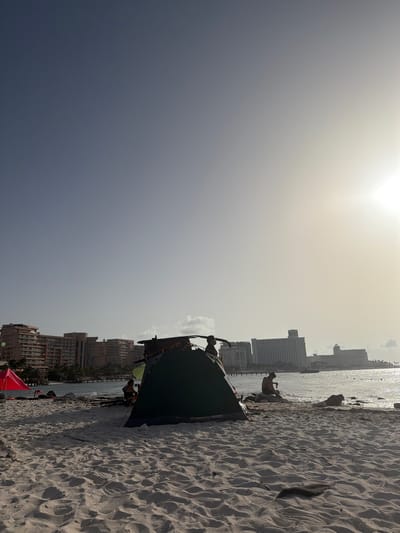 The image depicts a beach scene in Cancun, Mexico. The foreground is dominated by white sand with footprints and debris, leading to the water's edge where several people are visible. A green tent is set up on the beach, with two individuals near it. To the left, a small red tent is also present. In the background, the ocean stretches out, with several large resort buildings lining the coastline. The sun is low in the sky, casting a bright glow and creating a hazy atmosphere. The sky transitions from a clear blue at the top to a lighter, more washed-out tone near the horizon. The overall impression is a relaxed, tropical vacation setting.