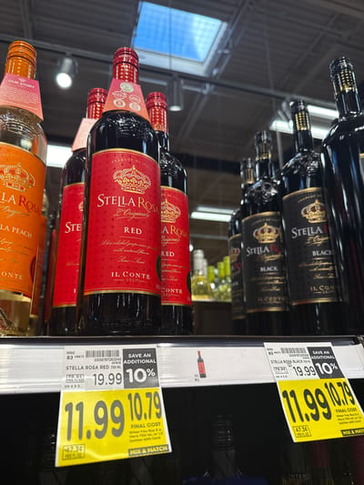 The image shows a shelf in a retail store, likely a grocery store or wine shop, in University Place, United States of America. The shelf is stocked with several bottles of Stella Rosa wine. In the foreground, there are two prominent price tags. One price tag is white and displays "STELLA ROSA RED 750 ML" with a price of "$19.99" and a "SAVE AN ADDITIONAL 10%" offer. Below this, a large yellow "MIX & MATCH" tag shows a final cost of "$11.99" with a separate price of "$10.79". The other visible price tag is similar, for "STELLA ROSA BLACK 750 ML," also indicating a "SAVE AN ADDITIONAL 10%" offer, with a regular price of "$19.99" and a final cost of "$11.99" and "$10.79". The bottles of wine are arranged on a white shelf. The lighting appears to be overhead retail lighting. The background shows more shelves stocked with similar wine bottles and a glimpse of a store ceiling with lights and ductwork, and possibly a skylight. There are no people visible in the image.