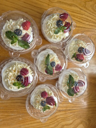 The image shows six individual servings of meringue desserts, each in a clear plastic container with a lid. The desserts are topped with whipped cream, fresh berries (raspberries and blueberries), slices of what appear to be strawberries or cherries, and mint leaves. Small golden sprinkles are scattered over the cream. The desserts are arranged on a wooden surface, likely a table, with visible wood grain. The lighting suggests it could be daytime, indoors, or outdoors with indirect light. The setting is a casual, possibly domestic, environment. There is no visible text. The desserts appear freshly made and appealing.