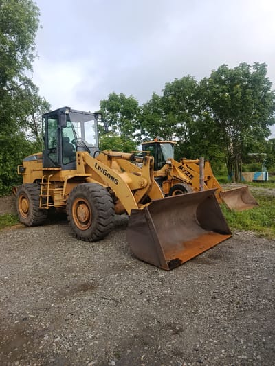 Two yellow wheel loaders are parked on a gravel surface in what appears to be an outdoor storage area. The loader in the foreground is a LIUGONG model, positioned facing slightly to the right. It has large, treaded tires and a substantial bucket in the lowered position. The cab is visible with dark tinted windows. Behind it, a second, similar wheel loader is partially visible, also yellow and with its bucket lowered. Trees with lush green foliage form a backdrop to the scene. The sky is overcast with a light gray hue. The ground is a mix of gravel and some patches of wetness, suggesting recent rain. There are no people or active operations depicted. The environment appears rural or semi-industrial, with some distant structures visible, possibly part of a facility or settlement, but no specific landmarks in Khorol, Russia, are discernible.