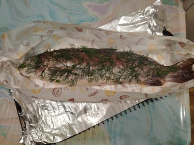 A whole fish, covered in dill, lies on a baking sheet lined with parchment paper and aluminum foil. The parchment paper has a pattern of small drawings, including croissants, candies, and suns. The fish is silver and red with a dark brown tail. The baking sheet is placed on a table with a blue and white marbled pattern. The lighting is bright, indicating an indoor setting. The image quality is clear, allowing for detailed observation of the fish and its surroundings. There are no people present in the image.