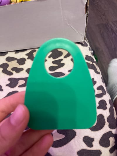 In the city of Vaganovo, Russia, a person's hand with visible fingernails is holding a bright green, plastic object with a rounded top and a circular cutout in the center. The object is positioned over a surface with a black and white cow-print pattern. In the background, a cardboard box is visible, with the edge of a colored item peeking out from inside. A person's foot wearing a gray sock is also visible on the right side of the frame. The overall setting appears to be indoors, with a wooden floor visible in the far background.