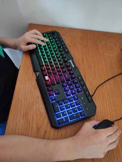 In Jablonec nad Nisou, Czechia, on a wooden desk, a person's hands are visible interacting with a black, RGB-backlit keyboard and a black mouse. The keyboard features illuminated keys in various colors, including green, pink, and blue. The person's left hand rests on the keys, and their right hand is positioned on the mouse. The scene is captured from an elevated perspective, looking down at the desk and the computing peripherals.