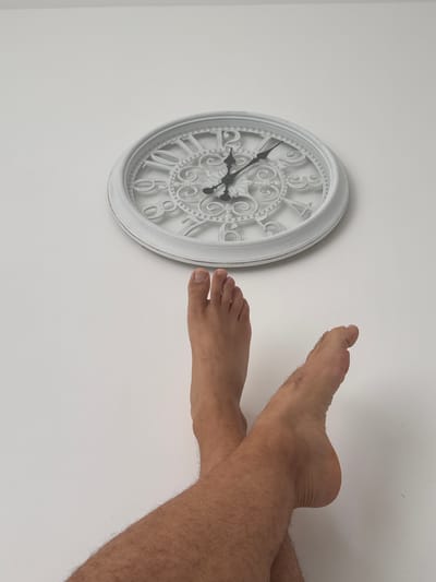 A white wall serves as the backdrop for an ornate white clock displaying Roman numerals. The clock's hands indicate it is approximately 5:13. Positioned below the clock, on the floor, are two bare feet belonging to a person. The feet are crossed at the ankles, with one foot turned slightly inwards. The skin on the feet and legs appears to be light-toned with visible hair. The scene is devoid of any discernible urban context, and no specific location within Molenbeek-Saint-Jean - Sint-Jans-Molenbeek, Belgium can be determined from the visual information provided.