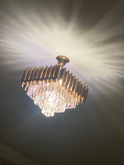 A modern chandelier is depicted hanging from a ceiling in Abad Tukimachi, Uzbekistan. The chandelier features a gold-toned metallic frame with a conical canopy affixed to the light-colored ceiling. Below the canopy, the frame is composed of numerous vertical, rectangular, gold-colored panels arranged in multiple tiers. Beneath these panels, several horizontal tiers of clear, rectangular, faceted crystal elements are suspended, actively emitting bright white light. This illumination casts a distinct radial, starburst-like pattern of light and shadow across the ceiling surface directly above the fixture. The lower portion of the image is dark, indicating the surrounding environment below the chandelier. No individuals, actions, or interactions are visible.
