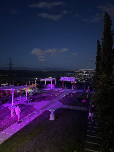 This image depicts an outdoor evening scene at a venue, likely a restaurant or event space, situated on a hillside overlooking a city at dusk. The sky is a deep navy blue with scattered, wispy clouds. In the foreground, a person is seen lying on a grassy area, illuminated by purple lighting. Surrounding them are outdoor furniture, tables, and umbrellas, also bathed in purple light, suggesting a festive or atmospheric setup. A pathway with decorative rope fencing runs through the scene.
Further into the background, illuminated structures and tables suggest seating areas and possibly a dance floor or lounge. The city lights of a sprawling urban area are visible in the distance, painting a warm glow against the darkening landscape. The presence of a large, metal electricity pylon on the left side of the image indicates a semi-rural or transitional zone between the city and the venue. The overall mood is relaxed and possibly celebratory, with the purple lighting creating a distinct ambiance. The time of day is clearly evening, as indicated by the twilight sky and the city lights. The weather appears clear.