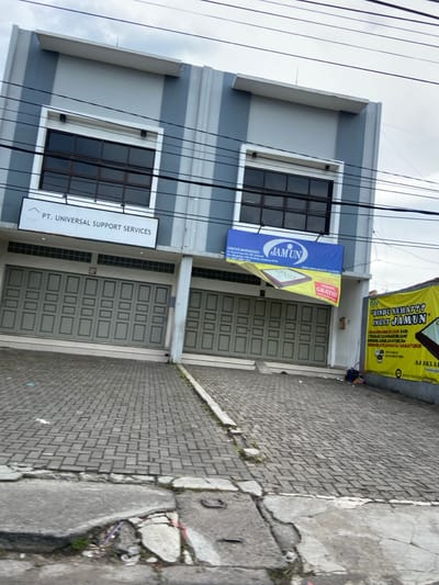 The image shows a street-level view of two commercial storefronts in Margahayu, Indonesia. The buildings are both constructed of concrete, with gray walls and gray tiled roofs. The storefronts have large, closed, green-painted wooden doors. The storefronts are located on a sloped paved driveway. The storefronts each have a sign above their doors, the first one reads "PT. UNIVERSAL SUPPORT SERVICES," and the second sign reads "CENTER MARGAHAYU JAM UN". The second storefront also has a larger sign, mostly yellow, that reads "RINDU SEHAT?? INGT JAMUN." The signs are visible against a bright sky with many power lines. A few plants and a small amount of trash are visible in the foreground.
