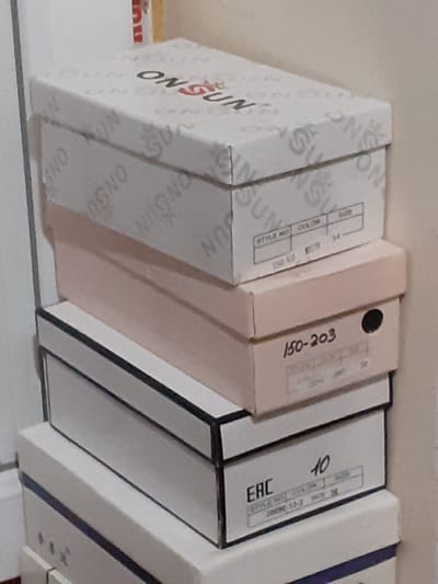This media file showcases a collection of stacked shoe boxes, likely in a retail or storage setting. The boxes are arranged on a shelf against a plain, light-colored wall. The primary subject matter consists of these cardboard boxes, many of which feature branding and labeling.
Several boxes display the prominent "ONSUN" logo in a repeating pattern. Other visible text includes "STYLE NO", "COLOR", and numerical identifiers such as "34", "150-203", and "10". One box also has the "EAC" mark. The colors of the boxes vary from white with black and red lettering to a pale pinkish-beige box. The perspective suggests a slightly upward angle, looking at the boxes from below. There are no people or clear indications of a specific time of day or weather conditions. The location context suggests this could be a store in Khujand, Tajikistan. The overall impression is one of organized inventory.