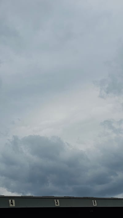 The image shows a view of a cloudy sky above a section of bleachers or a stadium grandstand. The top edge of the grandstand is visible, and on it are white numbers on black squares. The visible numbers are "4", "5", and "6", indicating seating sections. The sky is filled with heavy, gray clouds, suggesting overcast or potentially rainy weather. The scene appears to be outdoors, and based on the presence of numbered seating, it is likely a public venue such as a sports stadium or arena. The specific location in Neu-Ulm, Germany, is not discernable from the image alone, but it depicts a common scene at such venues. There are no people or specific actions depicted.