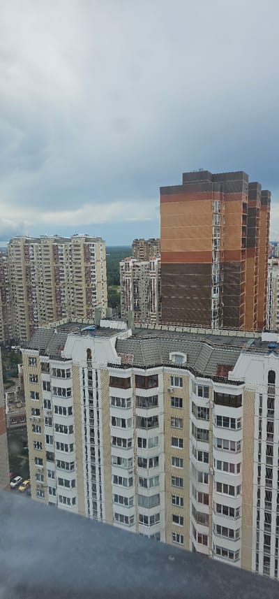 This is a vertical, eye-level shot of several tall, multi-story buildings in Butovo, Russia. The buildings are constructed in a modern, functional style, with repetitive balconies and windows. The closest building on the right is light yellow with white trim and dark windows, featuring numerous balconies that are set back from the facade. Further to the left, a darker, reddish-brown building with lighter brown stripes rises, appearing to be under construction or recently completed, as some windows are boarded up and construction materials may be visible. Beyond these, more buildings are visible in the background, receding into the distance under a hazy, overcast sky. The scene suggests an urban residential area. There is no visible text. The time of day appears to be daytime, given the ambient light, but the overcast sky obscures any strong shadows. The weather seems to be mild, as some windows are open and there's no indication of rain or extreme conditions.