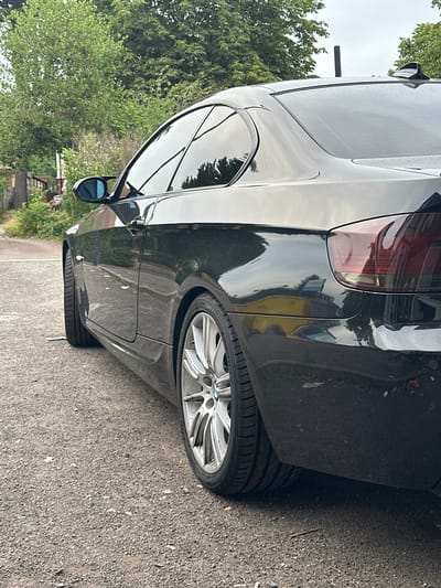 A black BMW coupe is parked on a gravel surface, its right side facing the camera. The car has tinted windows and a shark fin antenna on the roof. In the background, there are trees and a glimpse of a building. The scene appears to be in a residential area of Birmingham, United Kingdom, given the style of housing and vegetation visible. The overall lighting suggests an overcast day.