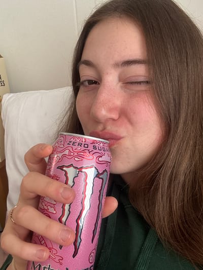 A young woman with brown hair and freckles is lying down, taking a sip from a pink Monster Energy Zero Sugar can. She is wearing a dark green top. The background consists of a white pillow and a blurry object that appears to be a cardboard box with text on it. The scene is indoors, likely in a bedroom. There are no discernible features that indicate the location is specifically in the city of Enschede, Netherlands.