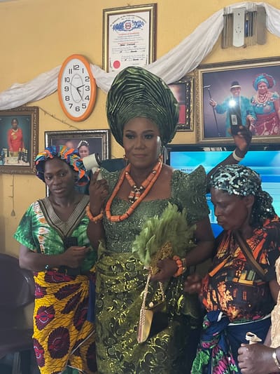 Here is a detailed description of the image:
**General Overview:**
The image captures a scene likely from a special occasion or event. It includes a woman dressed in elaborate traditional attire, possibly a bride or a celebrant, being attended to by two other women. The setting appears to be indoors, with walls adorned with pictures and decorations.
**People:**
1. **Woman in Green:** The focal point of the image is a woman wearing a green outfit with intricate beadwork and an elaborate headwrap of the same color. She is adorned with coral beads around her neck and wrist and holding a feathery fan.
2. **Woman Attending (Head Scarf):** An older woman wearing a headscarf with sequins is seen adjusting something on the main woman.
3. **Woman Attending (Head tie):** Another woman, with her head tied up in a blue and orange head tie, appears to be helping with the attire, possibly adjusting the lower part of the dress or gown.
**Objects and Environment:**
* The wall behind the women is decorated with framed pictures, certificates, and wall decor.
* An oval-shaped clock with a white face and orange frame is visible on the wall, showing the time at approximately 2:30.
* A length of white fabric is draped, adding to the decorative setting.
* A television screen is partially visible in the background, with the word "MUSIC" partially visible.
**Actions and Interactions:**
* The primary action is the assistance being given to the woman in the green outfit. The other two women are helping her, which suggests she is either getting ready for an event or having her outfit adjusted during one.
* The women are interacting closely, indicating care and attention to detail.
**Inferred Location (Warri, Nigeria):**
Given the traditional attire, cultural elements, and context clues, the setting is likely in Nigeria. Further information on the specific location (Warri) could be obtained from additional context about the cultural significance of the attire, decorations, and event in the image.