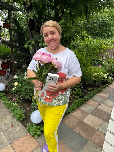 In the city of Nizhne-Chufichevo, Russia, a woman with short blonde hair stands in a garden setting, holding a bouquet of pink peonies and a silver iPhone 14 Pro Max in its box. She is wearing a white t-shirt with a floral and poppy print and bright yellow sweatpants with a white stripe. The garden features lush green trees and bushes, with a paved walkway made of square tiles in earthy tones. Several white spherical garden lights are visible, along with potted plants and decorative garden features. The woman is smiling and looking at the camera.