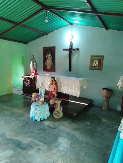 The image depicts the interior of a small chapel, likely located in a rural area, possibly Los Leoncitos, Venezuela. The chapel has a simple green corrugated metal roof supported by dark wooden beams and light green walls. A high altar is draped with a white cloth and adorned with religious statuettes of Mary and Jesus, a framed religious painting of Jesus and a dark wood crucifix. A small, round table covered with a light blue crocheted cloth holds a vase of flowers between statuettes of Jesus and Mary. The floor is of bare, unfinished concrete. A doorway to the left leads to another room. An ornate stone vase stands to the right of the altar. The scene is illuminated by sunlight streaming through a window behind the crucifix and a single bare light bulb hanging from the ceiling. Overall, the scene conveys a sense of humble devotion.