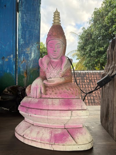 A weathered statue of Buddha, with peeling pink paint, sits on a wooden table. The statue is ornate, with a tiered headdress and intricate carvings on its robe and base. Behind the statue, a rustic blue wooden structure is visible, juxtaposed against a backdrop of green trees and a cloudy sky. To the right, a weathered wooden beam supports a black electrical cord with an on/off switch. The scene appears to be outdoors in a tropical environment, possibly in the city of Tejakula, Indonesia, given the architectural style of the tiled roof visible in the distance.
