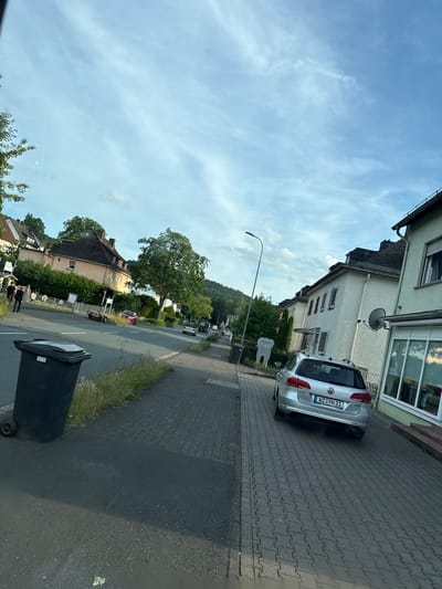 The image shows a street scene in Wetzlar, Germany. The foreground features a black trash bin on the left, positioned on a paved area next to a grassy verge. A silver Volkswagen Passat is parked on the right side of the street, facing away from the camera. The car has a German license plate that reads "WZ YN 21". Beyond the car, several residential buildings are visible, characterized by white and light-colored facades and dark-tiled roofs. A street lamp stands near the left side of the frame, and a large tree is prominent in the middle ground. The sky is partly cloudy with a bright blue hue. In the distance, a wooded hillside can be seen. Two figures, appearing to be young men, are walking on the sidewalk in the background on the left side of the image.