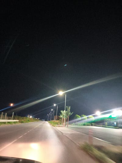 The image depicts a road at night, likely a highway or a major thoroughfare, as evidenced by the streetlights and the width of the road. The scene is illuminated by artificial lights from streetlamps and possibly vehicle headlights, casting long reflections on the wet or dark asphalt. On the right side of the road, there are several streetlights, some with bright, elongated light flares due to the camera's exposure. Trees, possibly palm trees, are visible along the roadside. On the left side, a white barrier or guardrail runs parallel to the road. The sky is dark, with a few faint, distant lights or objects that are difficult to identify. The overall impression is a still, nocturnal urban or suburban landscape. There are no people or vehicles clearly visible or in focus, except for the blurred reflection of headlights in the foreground, suggesting the image was taken from a moving vehicle. The presence of streetlights and a paved road indicates an infrastructure commonly found in developed areas. Without additional information or context, it is not possible to definitively identify the specific location within Yambol, Bulgaria, but it is consistent with roads found in such a city.