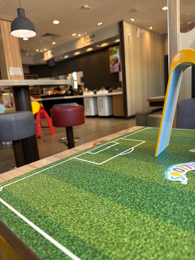This image shows a close-up of a table with a printed soccer field design. A yellow and blue object, possibly a toy goalpost, is positioned on the right side of the soccer field. The background is blurred, but appears to be an indoor restaurant setting, likely a McDonald's, based on the signage that reads "CASSA" (Italian for "cashier" or "till") and what looks like a promotional poster for a burger. The restaurant appears to be located in Domodossola, Italy, as suggested by the presence of Italian text. Other elements visible in the background include tables, chairs, and ceiling lights. The overall lighting suggests it is daytime. There are no people clearly visible or engaged in any specific actions in the foreground.