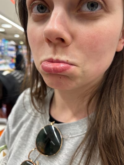 A close-up, chest-up indoor photograph captures a female individual with long brown hair, pale complexion, and light blue eyes. Her lips are pursed in a pout, appearing glossy and light pink, with a small brownish mark visible above the upper lip. She is dressed in a grey crew-neck sweatshirt, under which a black garment is visible at the neckline. Aviator-style sunglasses with gold-toned frames and dark lenses are hanging from the front of her sweatshirt. The background is blurred, showing the interior of a retail environment, likely a supermarket or pharmacy, characterized by shelves stocked with various products and bright overhead lighting. A blurred human figure in dark attire is discernible in the distant left background. The scene is situated in the city of Monsummano Terme, Italy.
