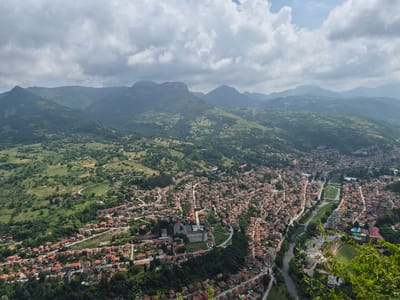 This image is an aerial view of Teteven, Bulgaria, on a cloudy day. The town is nestled in a valley surrounded by rolling green hills and mountains. The buildings are predominantly red-roofed houses, with a larger, lighter-colored building appearing to be a public institution or hotel in the center of the town. A river runs through the town, and there are roads winding through the residential areas. A green field, possibly a sports complex, is visible near the river. The foreground shows some lush green foliage, suggesting the vantage point is from a higher elevation overlooking the town. There are no visible people or specific actions taking place. The overall impression is a picturesque town situated within a natural landscape.