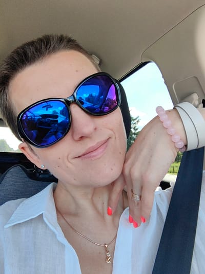 A close-up, selfie-style shot captures a person from the chest up, seated in the passenger seat of a vehicle. The person, wearing large, dark sunglasses with blue lenses that reflect the car's interior and the outside scenery, is looking towards the camera. They have short, dark hair, and are wearing a white collared shirt. A thin, rose gold necklace with a cat-shaped pendant is visible on their neck. On their right hand, which is resting on their cheek, they wear a rose-tinted beaded bracelet and a ring. Their fingernails are painted a bright coral color. The background includes the car's interior, specifically the ceiling and a seatbelt, as well as a glimpse of greenery and a bright sky through the car window. The location is not identifiable as Ilyanskiy selskiy Sovet, Belarus, or any other specific city, based solely on the visual information provided in the image.