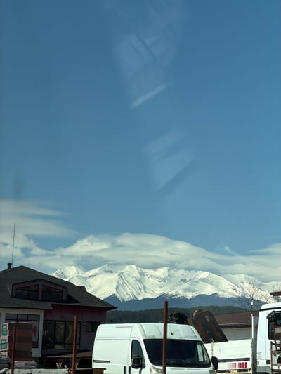 The image shows a view of snow-capped mountains partially obscured by clouds under a blue sky. In the foreground, on the right side, there are buildings with dark grey roofs and white and red facades. A white van is parked near the building, and a large truck is parked slightly behind the van. Some metallic structures are present near the vehicles. The location is Kraishte, Bulgaria.