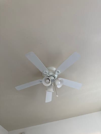 A white ceiling fan with three blades is mounted on a textured, off-white ceiling. The fan has a central white housing with two light fixtures, each containing a white lampshade and a visible light bulb. A pull chain hangs from the center of the fan housing, with a white circular handle at the end. The fan is not in motion. The scene is indoors and appears to be a residential or office space. There are no people or animals visible. The lighting is ambient, suggesting the room is naturally lit or illuminated by the fan's bulbs. The location is in Glendale, United States of America.