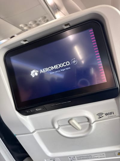 An in-flight entertainment screen shows the Aeromexico logo. The screen displays "Estar cerca. Llegar lejos." The device is encased in a white plastic console with a Wi-Fi sticker, a recline lever, a USB port, and two audio jacks. The console is attached to the back of an airplane seat. The lighting is bright, and the image is shot at a slight angle.