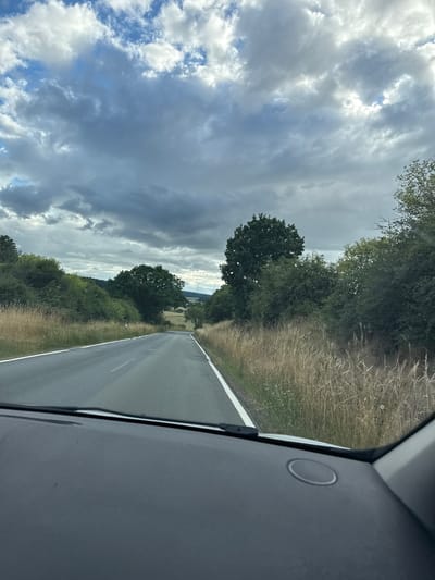 This is a vertical, first-person perspective shot from inside a vehicle, likely a car, traveling on a rural road in Oberwetz, Germany. The road stretches ahead, bordered by lush green trees and tall, dry grass on either side. The sky is dramatic and filled with a mix of dark, heavy clouds and patches of bright blue sky with fluffy white clouds, suggesting a dynamic and potentially changing weather. The overall impression of the weather is overcast with intermittent sun. The scene appears to be set during the daytime, given the visibility.
In the foreground, the interior of the vehicle is visible, specifically the dark gray dashboard on the right side of the frame, with a circular speaker grille. The road itself is asphalt and appears wet, indicating recent rain or dew. The trees lining the road are dense and varied in shape and shade of green, with some appearing more mature and broad-leafed than others. The tall, dry grass to the right of the road suggests a countryside setting, possibly late summer or early autumn. In the distance, the road curves slightly, leading towards a gently rolling landscape with hints of more trees and open fields. There are no people or animals visible in the frame. The overall mood is one of travel through a scenic, somewhat moody landscape. No text is visible in the image.